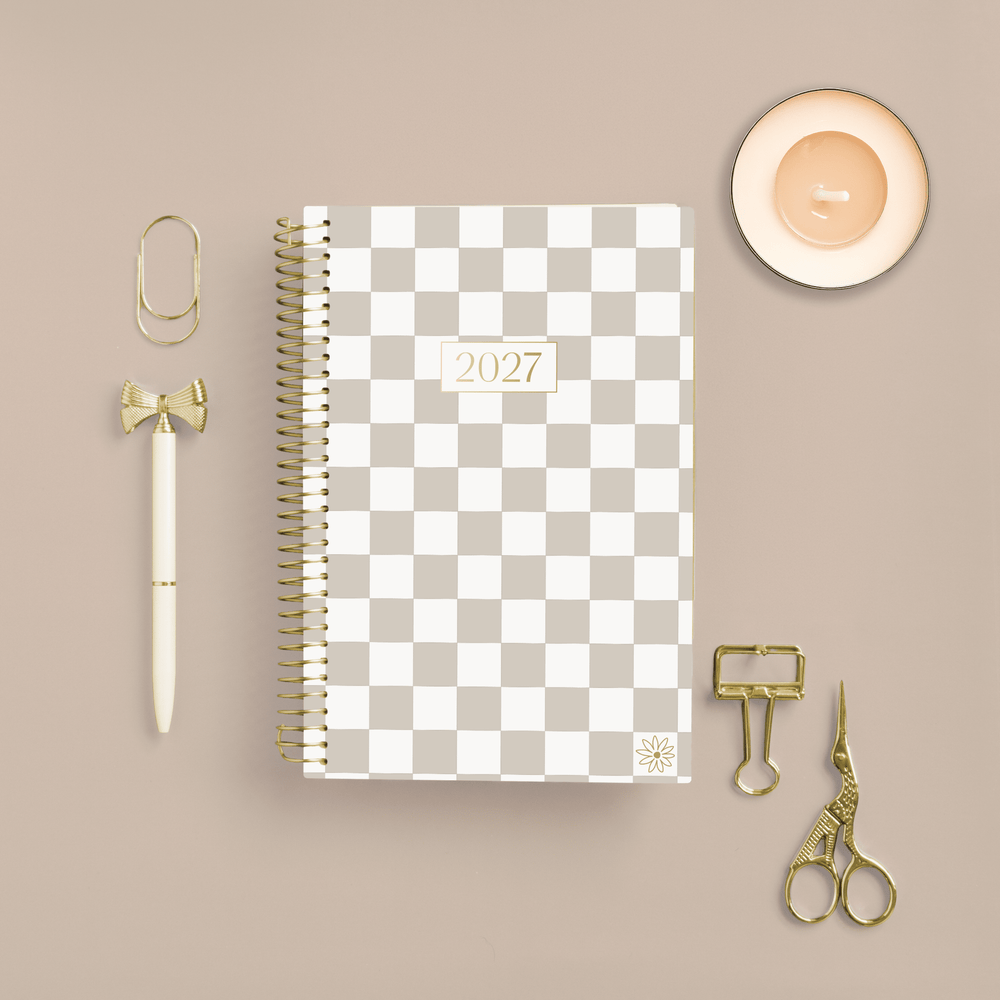 A 2027 spiral-bound planner with a beige and white checkerboard cover is displayed in a styled flat-lay arrangement on a warm beige background, surrounded by gold and cream stationery accessories including a pen, paperclips, binder clips, and scissors. - hover