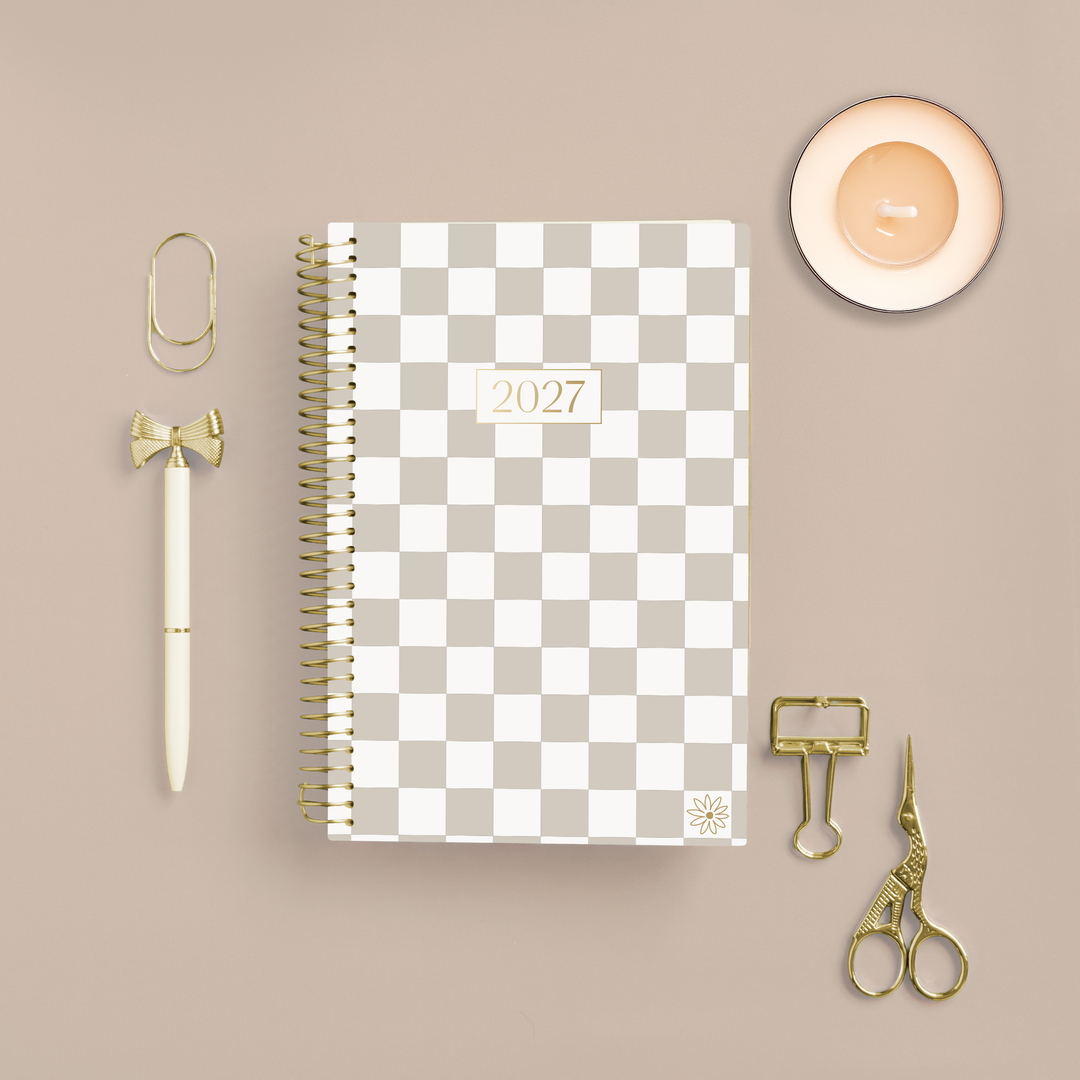 A 2027 spiral-bound planner with a beige and white checkerboard cover is displayed in a styled flat-lay arrangement on a warm beige background, surrounded by gold and cream stationery accessories including a pen, paperclips, binder clips, and scissors.