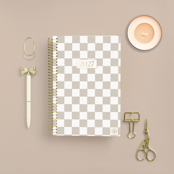 A 2027 spiral-bound planner with a beige and white checkerboard cover is displayed in a styled flat-lay arrangement on a warm beige background, surrounded by gold and cream stationery accessories including a pen, paperclips, binder clips, and scissors.