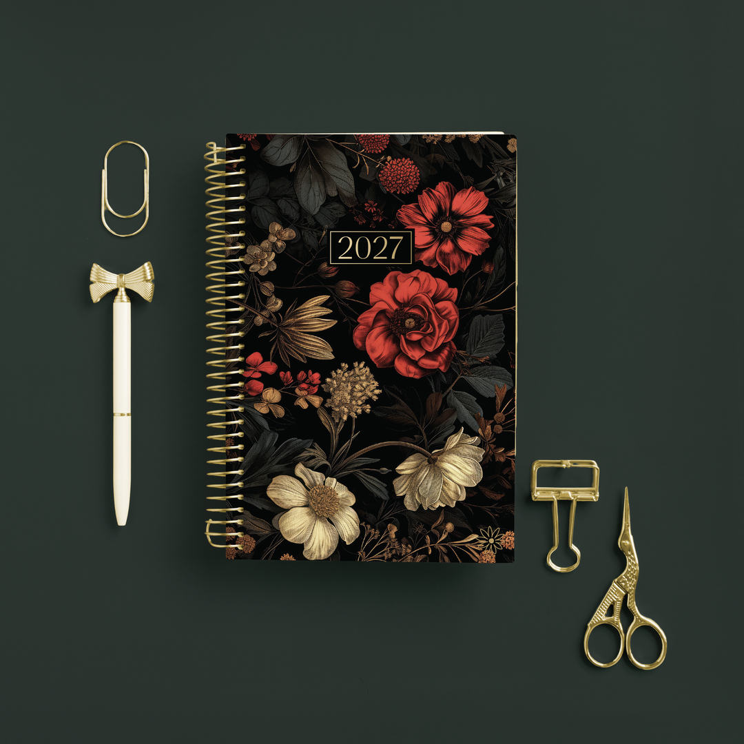 A 2027 spiral-bound planner with a dark floral cover, styled in a flat-lay arrangement on a dark green background alongside gold stationery accessories including a paperclip, pen, binder clips, and scissors.