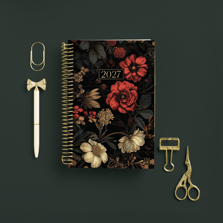 A 2027 spiral-bound planner with a dark floral cover, styled in a flat-lay arrangement on a dark green background alongside gold stationery accessories including a paperclip, pen, binder clips, and scissors.