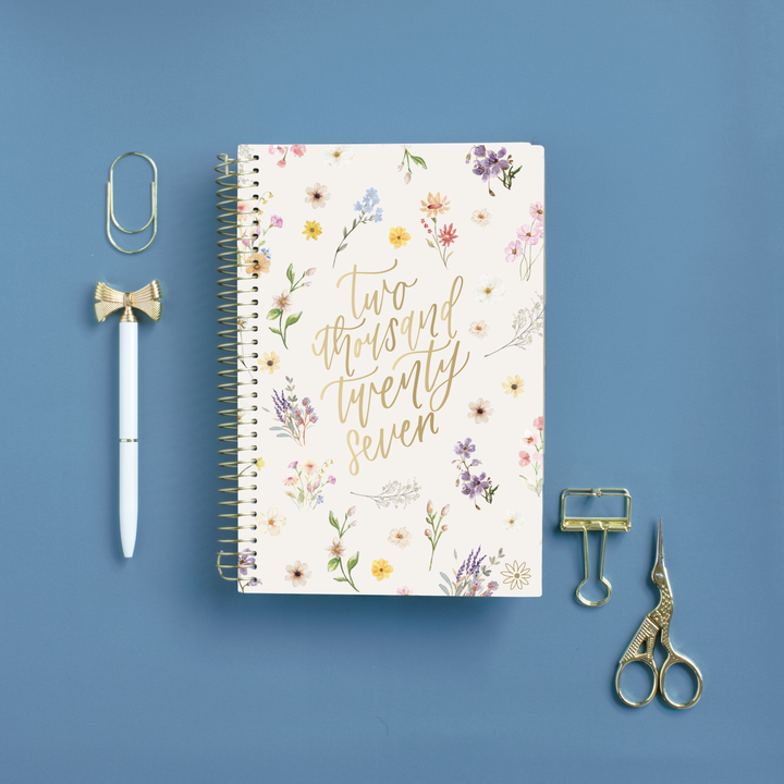 A spiral-bound notebook with a cream floral cover and gold script reading 'Two thousand twenty seven' is arranged on a blue background with stationery props including a paperclip, pen, binder clip, and scissors.