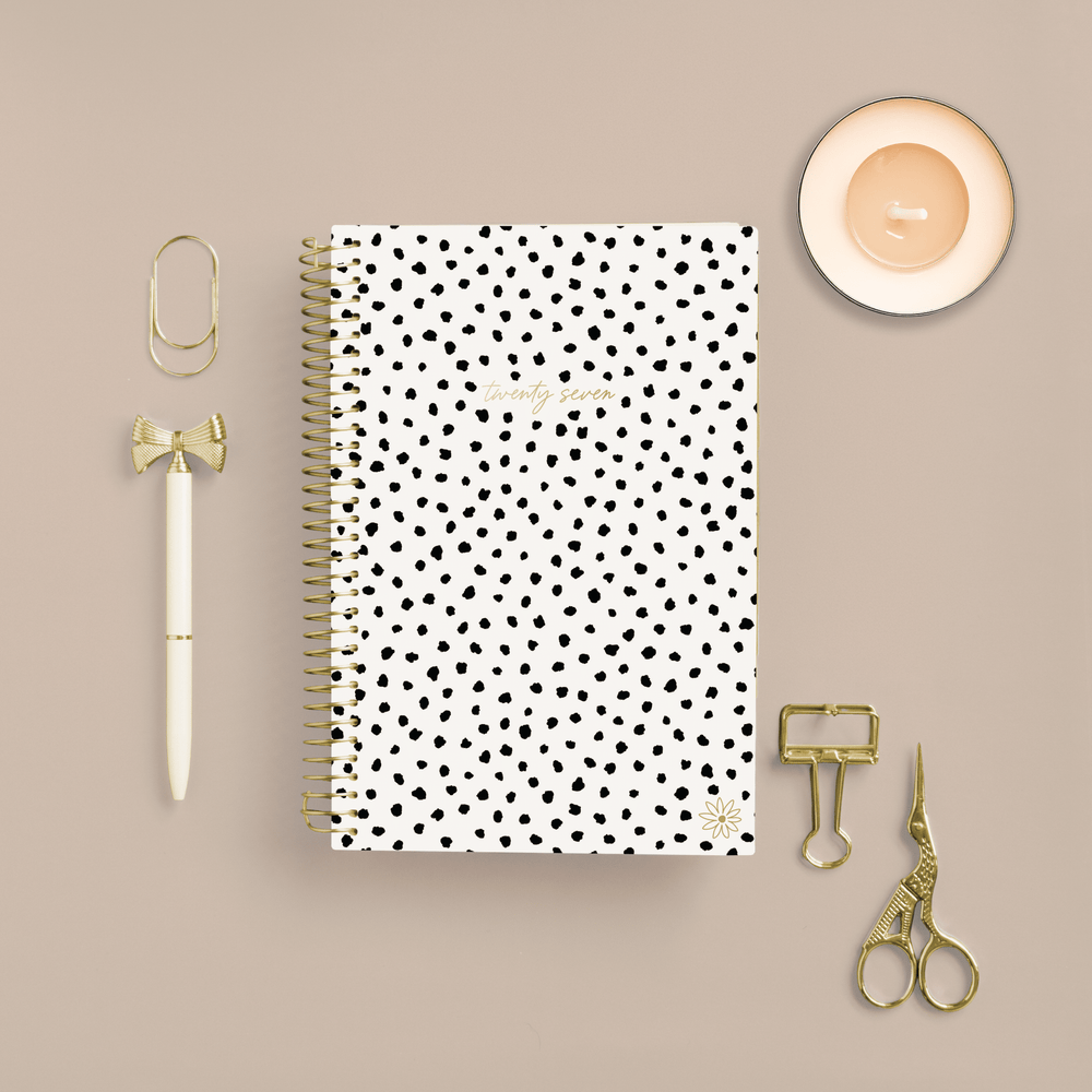 A spiral-bound planner with a white cover and black polka dots is arranged in a flat lay on a beige background, surrounded by gold stationery accessories including a pen, paperclips, scissors, and a small candle. - hover