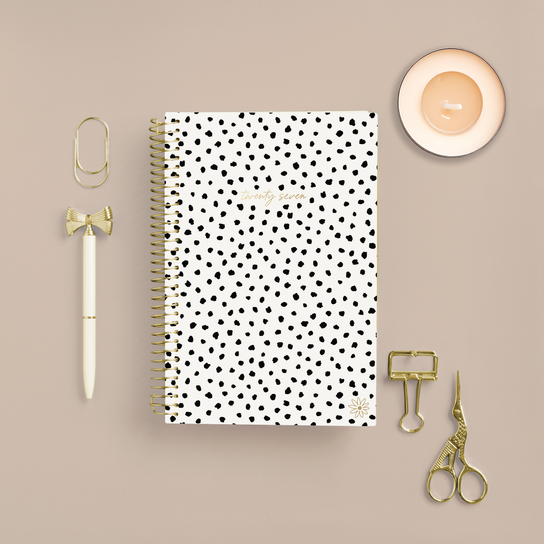 A spiral-bound planner with a white cover and black polka dots is arranged in a flat lay on a beige background, surrounded by gold stationery accessories including a pen, paperclips, scissors, and a small candle.