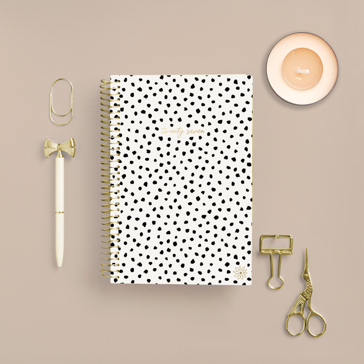 A spiral-bound planner with a white cover and black polka dots is arranged in a flat lay on a beige background, surrounded by gold stationery accessories including a pen, paperclips, scissors, and a small candle.
