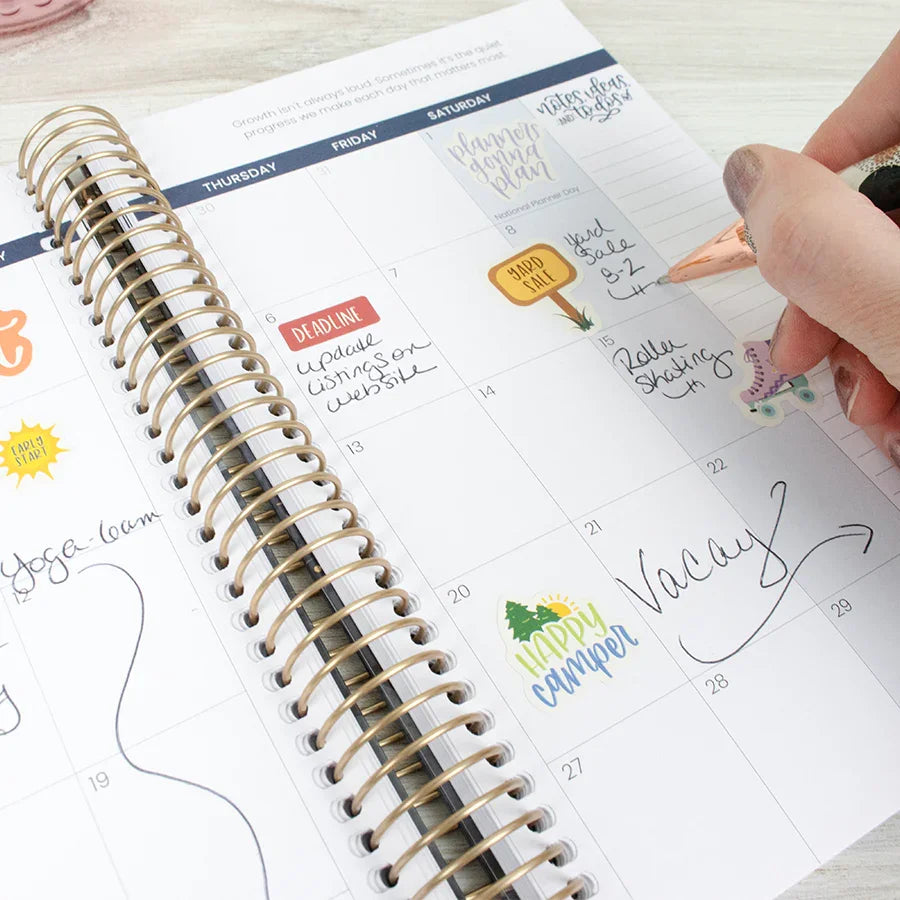 A hand writes in a bloom daily planners 2026 Hard Cover Planner, 5.5" x 8.25", Midnight Sky, open to the monthly calendar with colorful stickers, notes like "Vacay," arrows, and decorative icons on the pages.