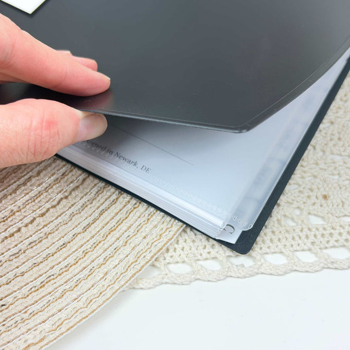 A close-up of a hand holding a black folder or binder open over a white surface, with a beige woven placemat and cream crochet fabric underneath.