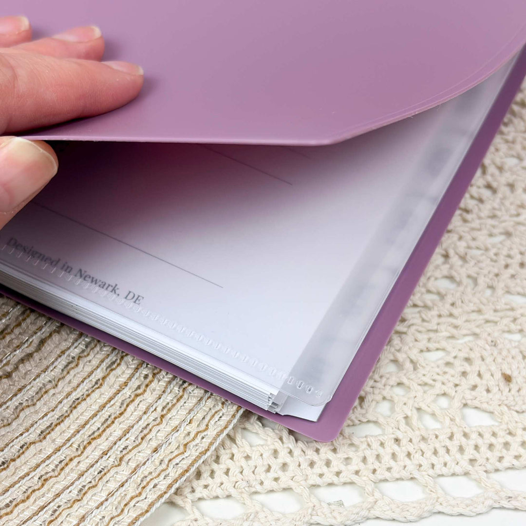 A close-up of a hand lifting the flap of a lavender purple file folder to reveal white document inserts inside, placed on a cream crocheted textile surface.