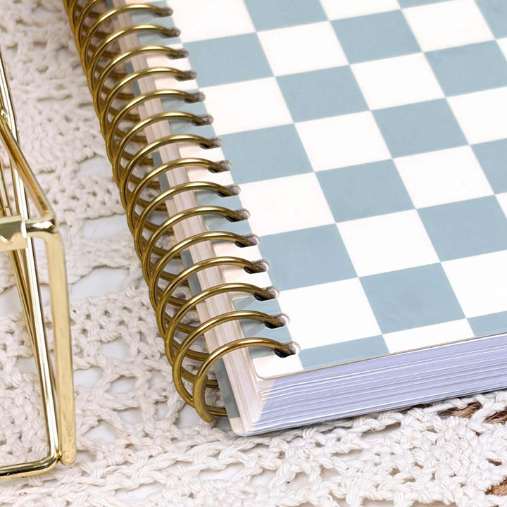 A spiral-bound planner with a light blue and white checkerboard cover, shown on a white lace doily next to a gold wire tray.