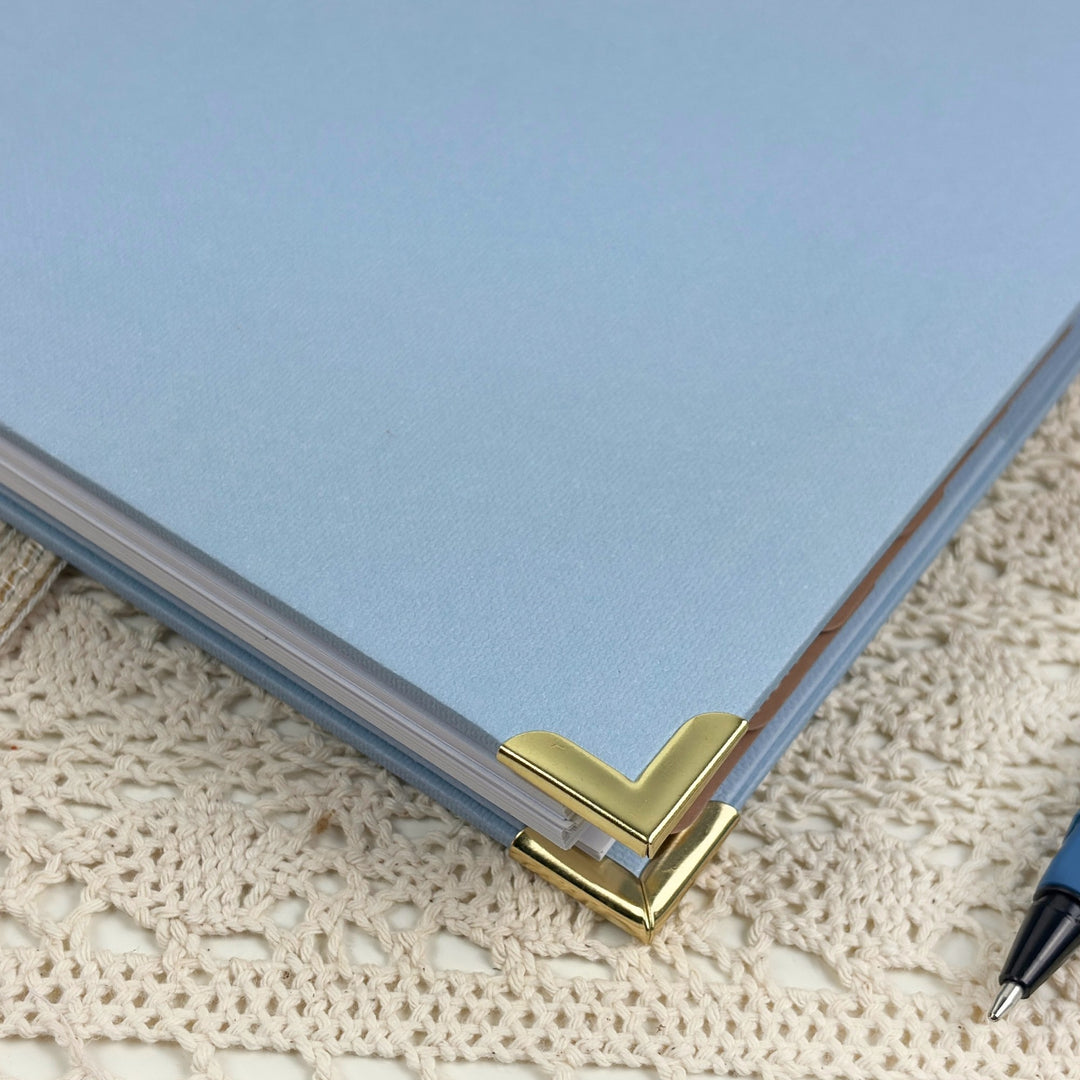 A close-up of a light blue notebook or folder corner with a gold metal chevron corner protector, resting on a cream-colored crocheted lace surface.