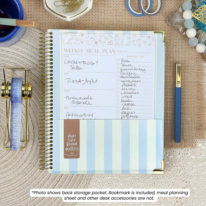 A spiral-bound weekly meal planner notebook shown open on a woven beige desk mat, surrounded by desk accessories including a blue pen, binder clips, paper clips, and a small bowl. The planner has a white page with handwritten notes, pale blue and white vertical stripes on the cover, and a small brown bookmark pocket.