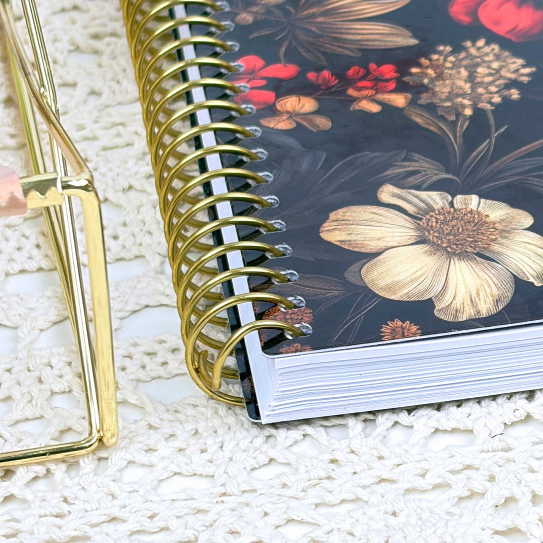 A close-up of a spiral-bound notebook with a dark floral cover, featuring a large cream flower and red blossoms, placed on a white lace surface next to a gold metal stand.