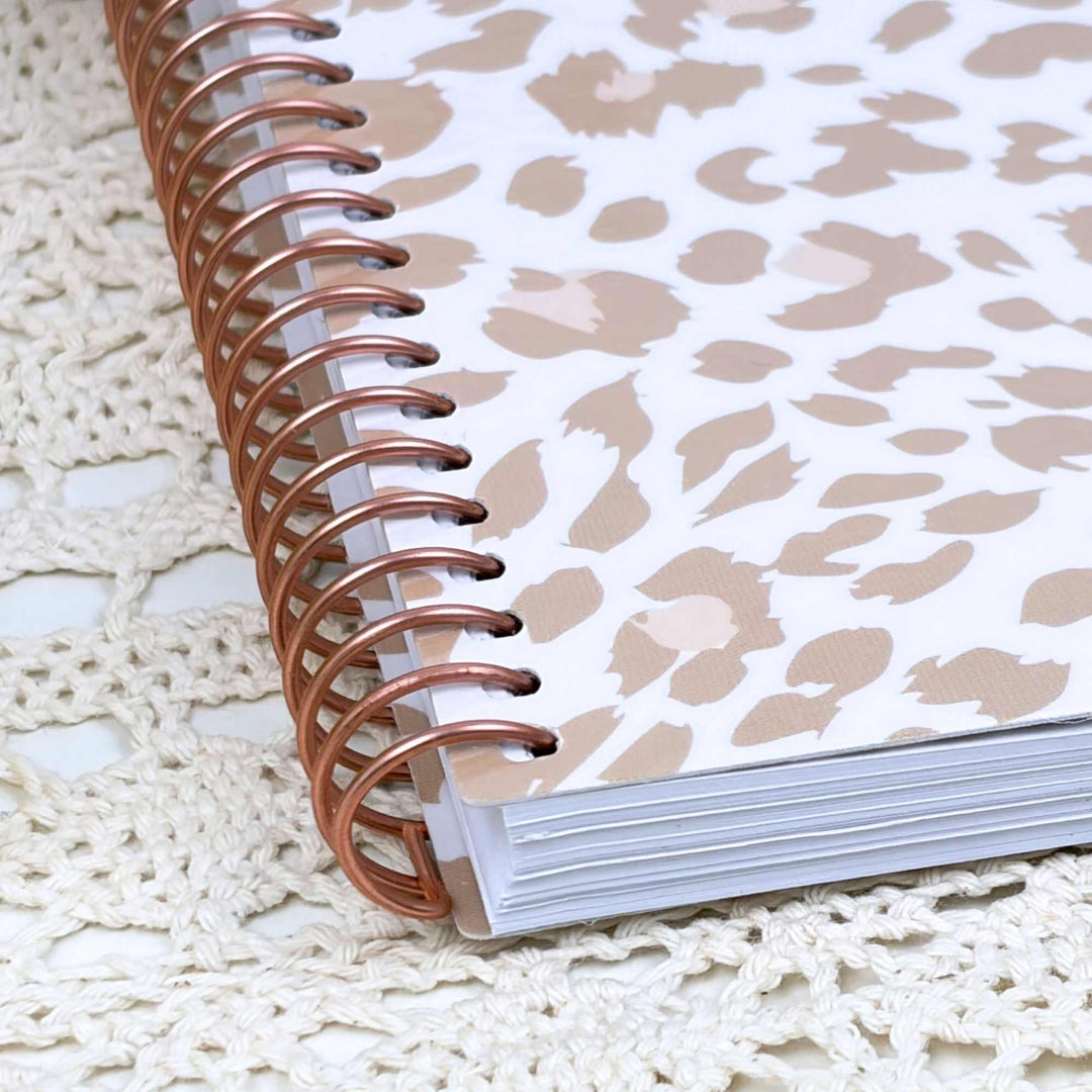 A spiral-bound notebook with a white cover featuring a beige and light pink leopard print, bound with metallic rose gold rings, resting on a cream-colored crocheted lace doily.