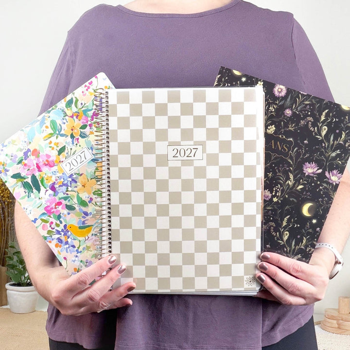A person in a purple top holds three spiral-bound 2027 planners. The center planner has a beige and white checkerboard pattern, while the left planner has a colorful floral design and the right planner is black with gold celestial illustrations.