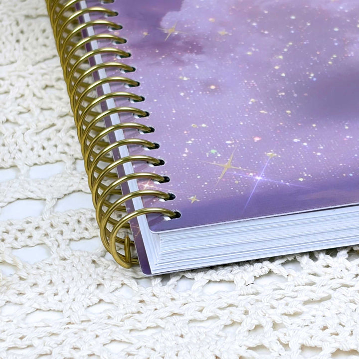 A spiral-bound notebook with a purple galaxy-patterned cover and shiny gold wire binding, placed on a white crocheted textile surface.