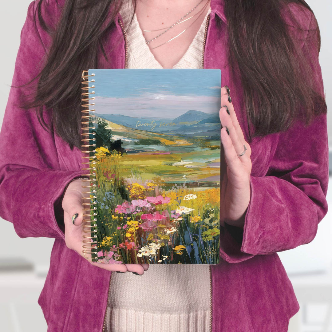 A person in a magenta velvet jacket holds a spiral-bound notebook featuring a colorful landscape painting of a wildflower meadow and distant mountains.