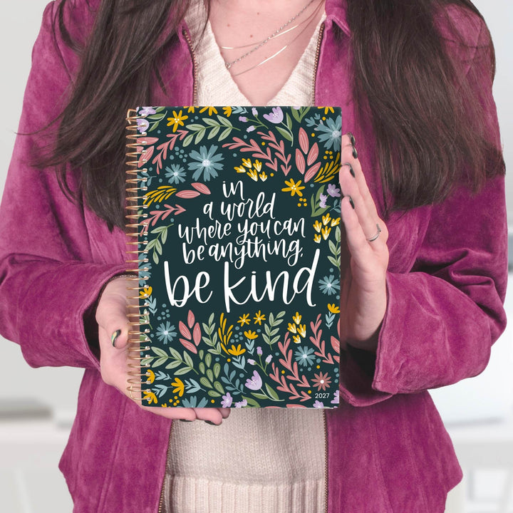 A person in a magenta velvet jacket holds a spiral-bound notebook with a floral pattern and the quote 'in a world where you can be anything be kind' on the cover.