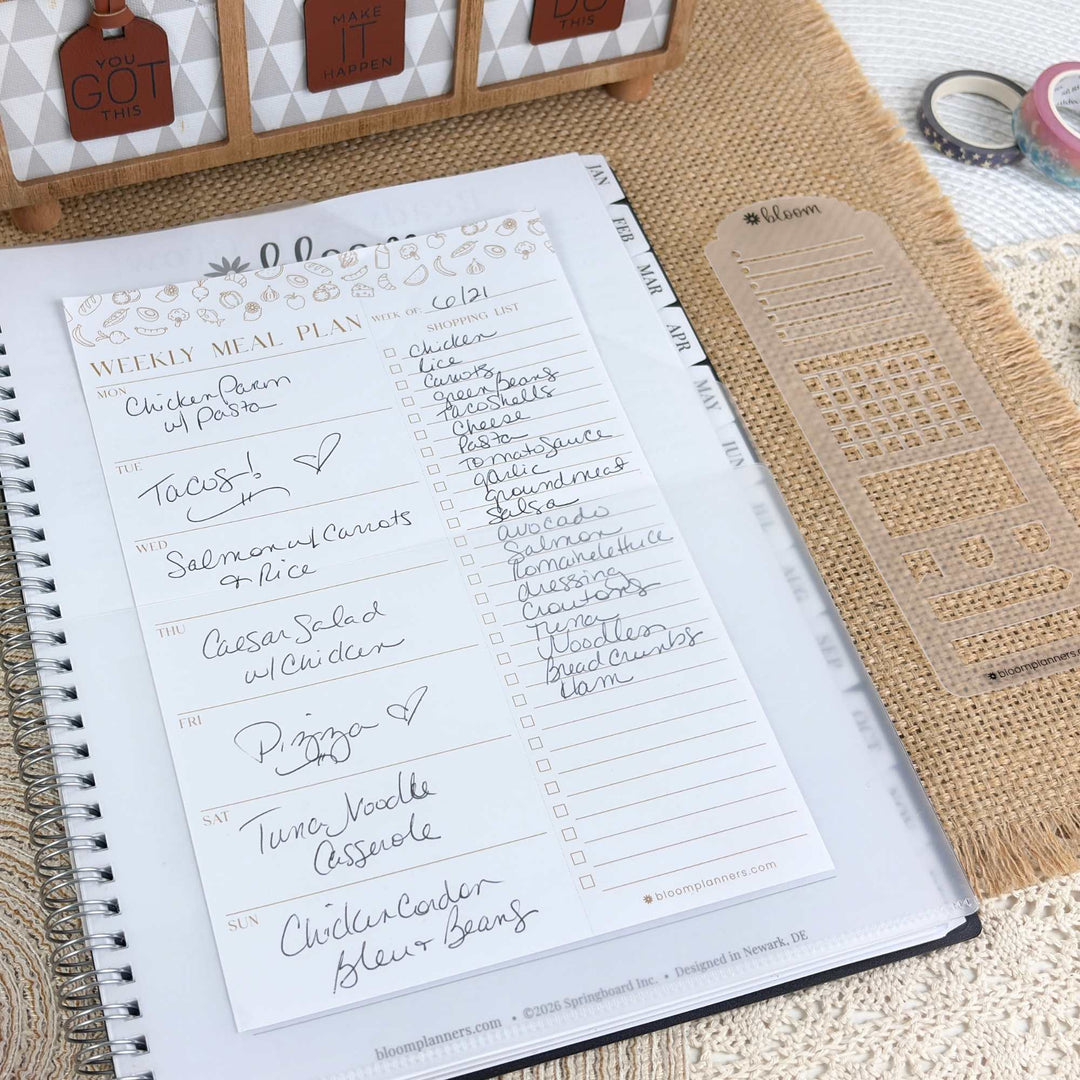 A weekly meal plan insert page from a spiral-bound planner, laid on a burlap surface with a clear plastic divider and decorative tags nearby.