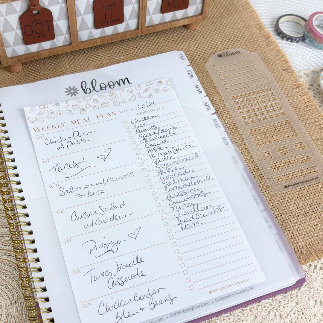 A weekly meal plan notepad from bloom planner, shown open in a spiral notebook with handwritten meal entries. A clear plastic divider page and decorative tags are placed beside it on a burlap surface.