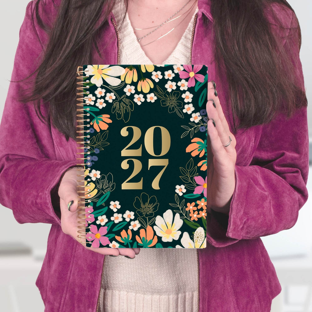 A person in a magenta velvet jacket holds a 2027 spiral-bound planner with a dark green floral cover. The planner features gold foil '2027' text surrounded by colorful flowers and green leaves. - hover