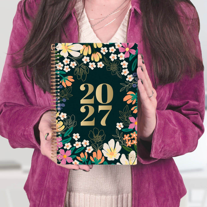 A person in a magenta velvet jacket holds a 2027 spiral-bound planner with a dark green floral cover. The planner features gold foil '2027' text surrounded by colorful flowers and green leaves.