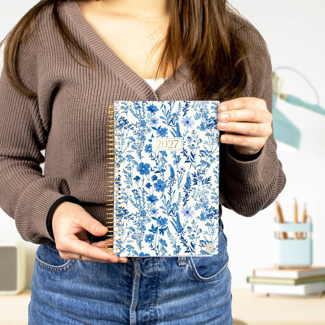 A person in a brown ribbed sweater and blue jeans holds a 2027 spiral-bound planner with a blue floral patterned cover in front of their torso. The background shows a softly blurred indoor desk area with a lamp and books.