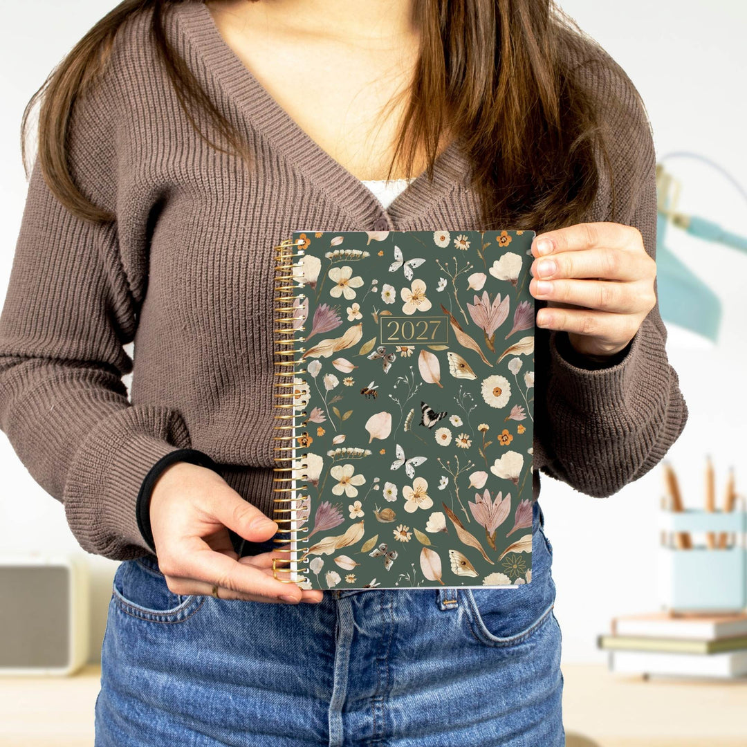 A person holding a 2027 spiral-bound planner with a dark green floral pattern cover, wearing a brown ribbed V-neck sweater and blue jeans.
