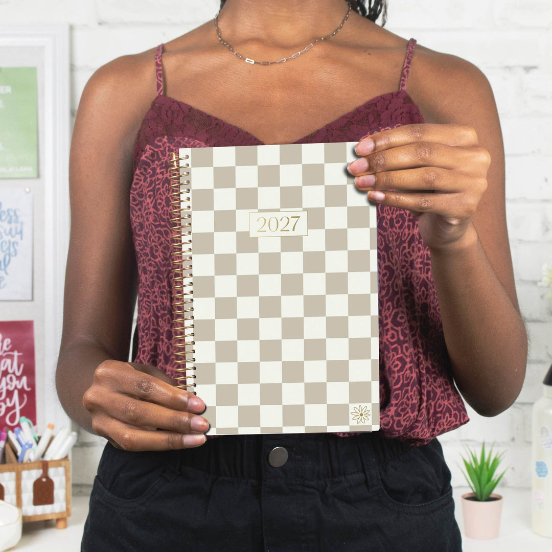 A person holds a 2027 spiral-bound planner with a beige and white checkerboard pattern. The person is wearing a burgundy patterned camisole top and black pants, standing in front of a white brick wall.
