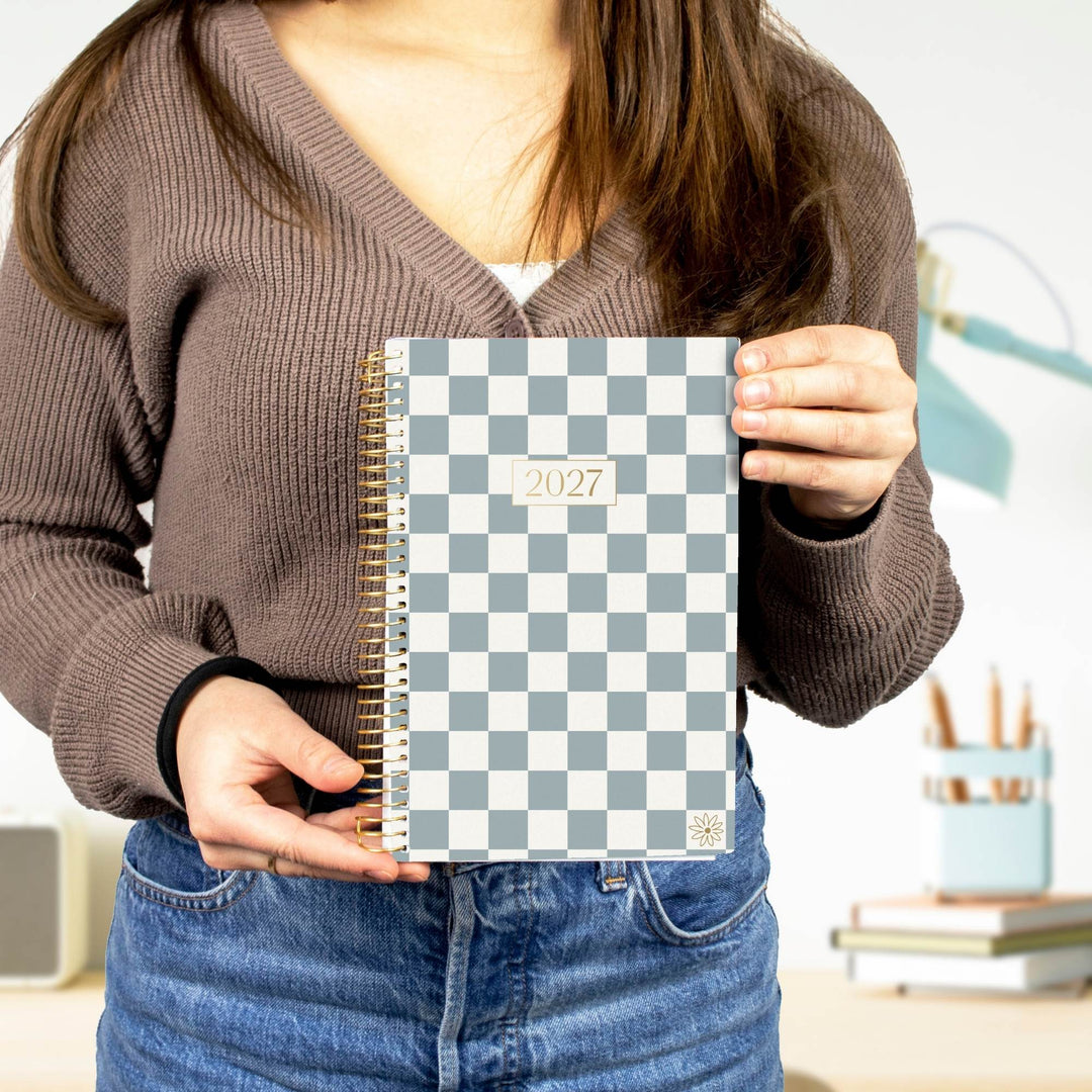 A person in a brown ribbed sweater and blue jeans holds a 2027 spiral-bound planner with a gray and white checkerboard cover in front of their torso.