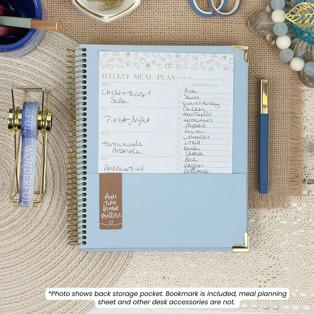 A spiral-bound weekly meal planner notebook in light blue with gold corner accents, shown open on a burlap surface with desk accessories around it. The page includes a printed header reading 'WEEKLY MEAL PLAN' and handwritten menu items.