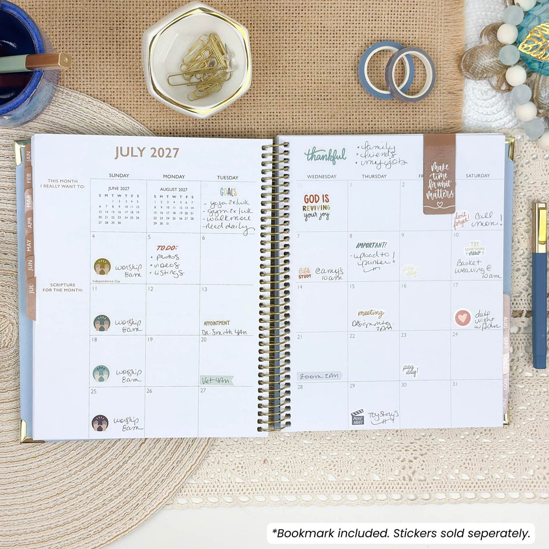 An open spiral-bound planner showing July 2027 with handwritten daily notes and stickers, styled on a woven beige surface with a blue mug, gold dish, washi tape, rings, and a pen around it.