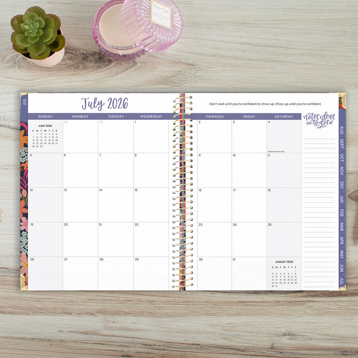 Open spiral-bound planner showing July 2026 on a wooden surface with a plant and candle.
