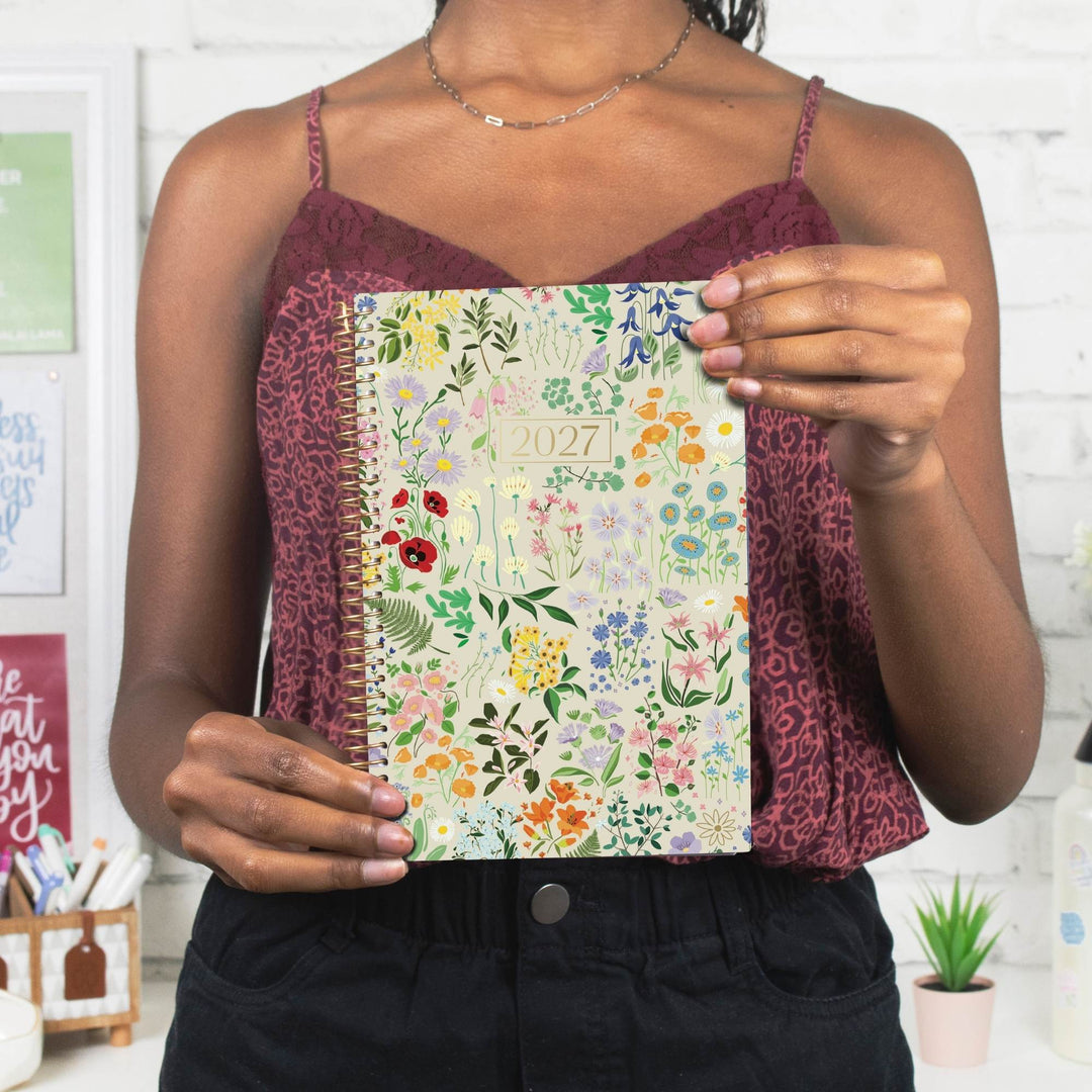 A woman holding a 2027 floral spiral-bound planner in front of her chest. She is wearing a burgundy patterned camisole and black pants, standing in a bright, white-themed room.