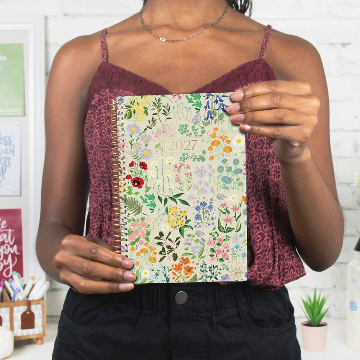 A woman holding a 2027 floral spiral-bound planner in front of her chest. She is wearing a burgundy patterned camisole and black pants, standing in a bright, white-themed room.