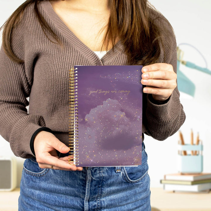 A person wearing a brown ribbed sweater and blue jeans holds a purple spiral notebook with a galaxy cloud design and gold foil text.