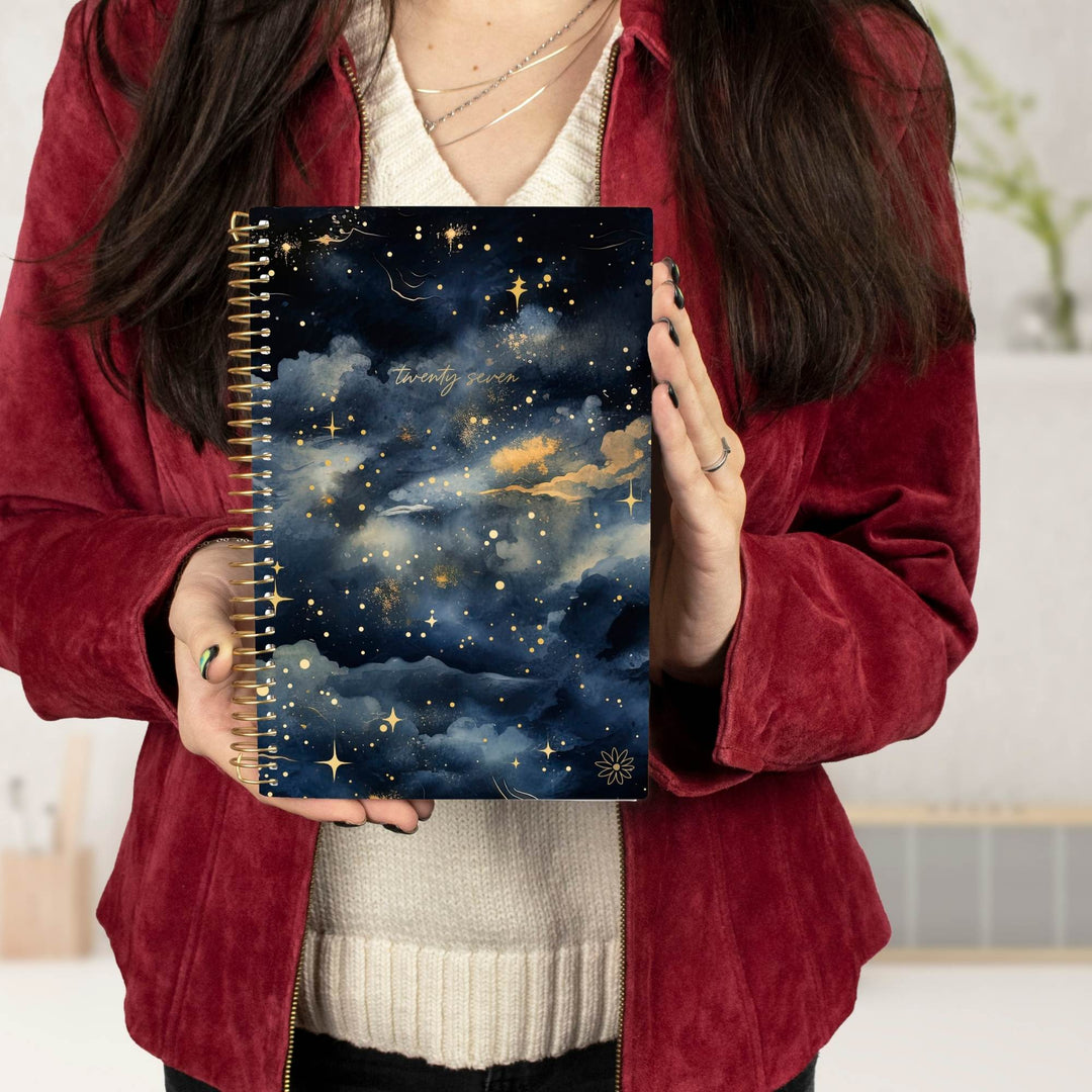 A person holding a spiral-bound notebook with a dark blue night sky cover featuring gold stars, clouds, and a crescent moon. The person is wearing a burgundy velvet jacket over a cream ribbed sweater.