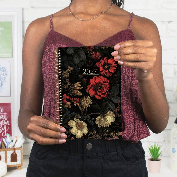 A woman holds a 2027 spiral-bound planner with a black floral cover featuring red, gold, and cream flowers. The planner is shown in a lifestyle setting, held up by a person wearing a burgundy patterned camisole and black pants.