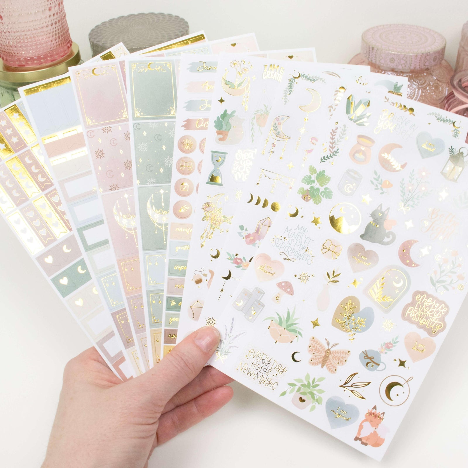 A hand holds the bloomdailyplanners Simply Gilded x bloom Boho Celestial Sticker Pack with pastel and gold foil moons, stars, plants, animals, and shapes; pink storage containers are in the background.