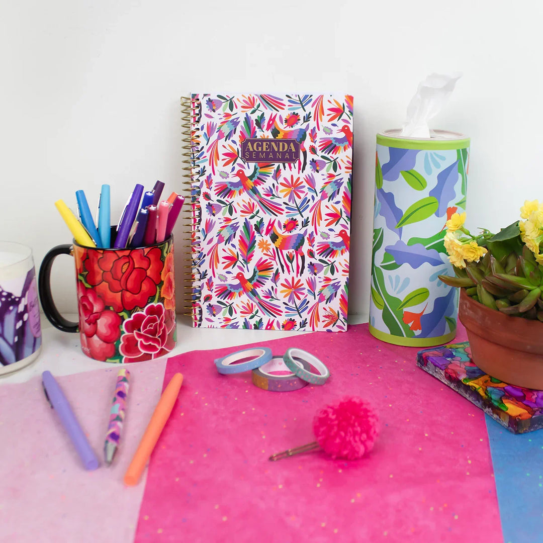 Colorful stationery items including a floral notebook, pens, and a cup on a pink surface.