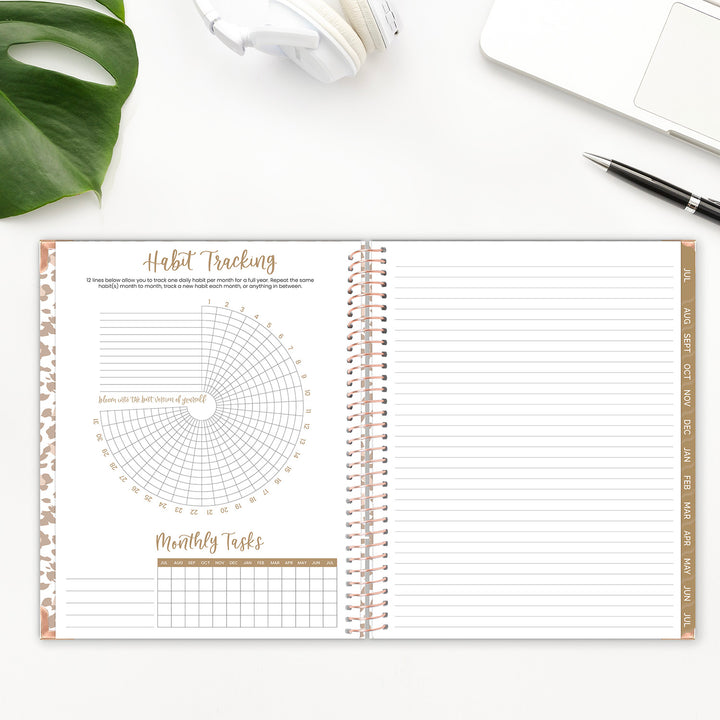Open spiral-bound planner with habit tracking and monthly tasks sections on a white surface with a plant and pen.