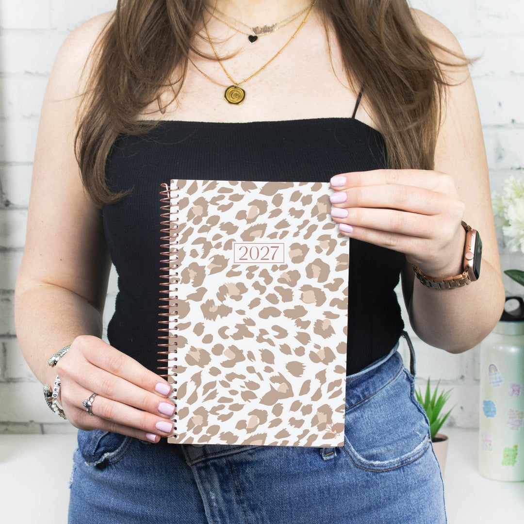 A woman in a black camisole and blue jeans holds a spiral-bound 2027 planner with a beige and brown leopard print cover in front of her chest.