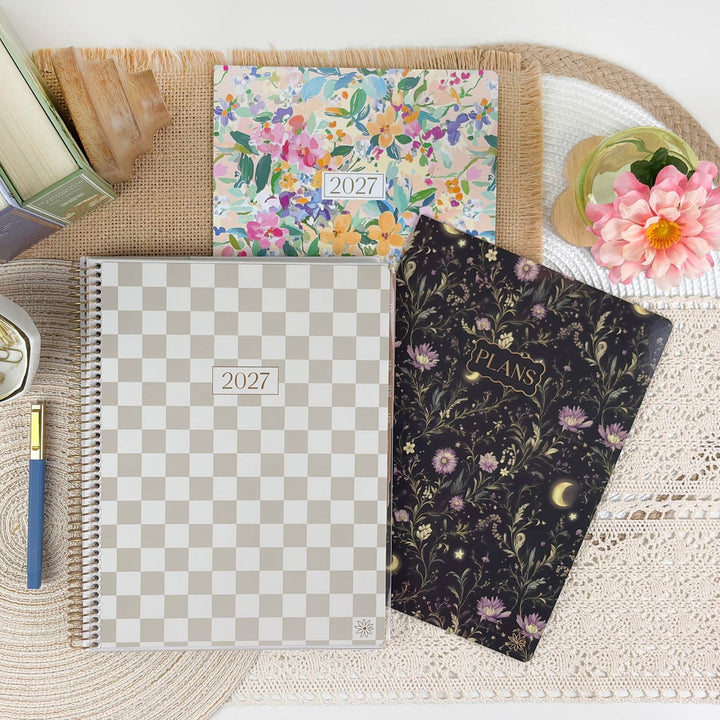 Three spiral-bound 2027 planners arranged on a woven placemat with decorative props. The front planner has a beige-and-white checkerboard cover with a small gold logo.