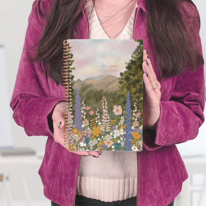 A person holding a spiral-bound notebook with a colorful floral and mountain landscape cover. The person is wearing a magenta corduroy jacket over a cream knit sweater.