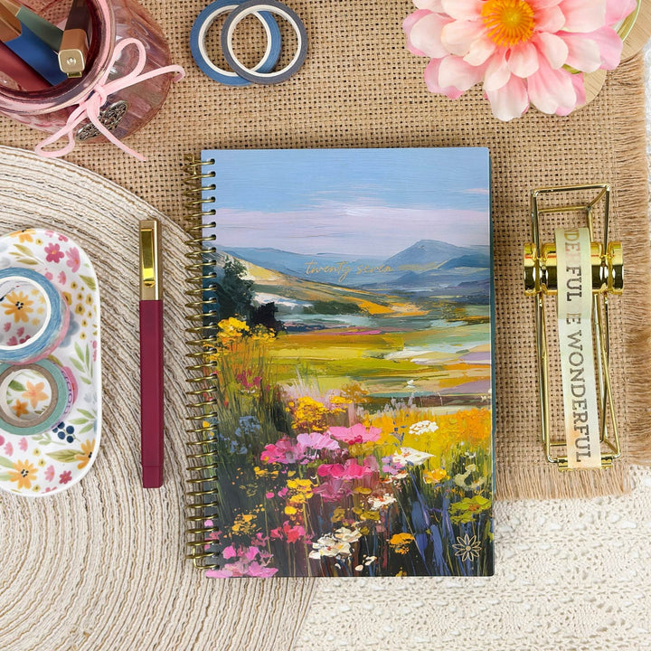 A spiral-bound notebook with a colorful landscape painting cover featuring wildflowers and mountains, styled in a flat-lay arrangement with a pink flower, gold binder clips, a floral pen, and other stationery on a burlap and woven mat background.