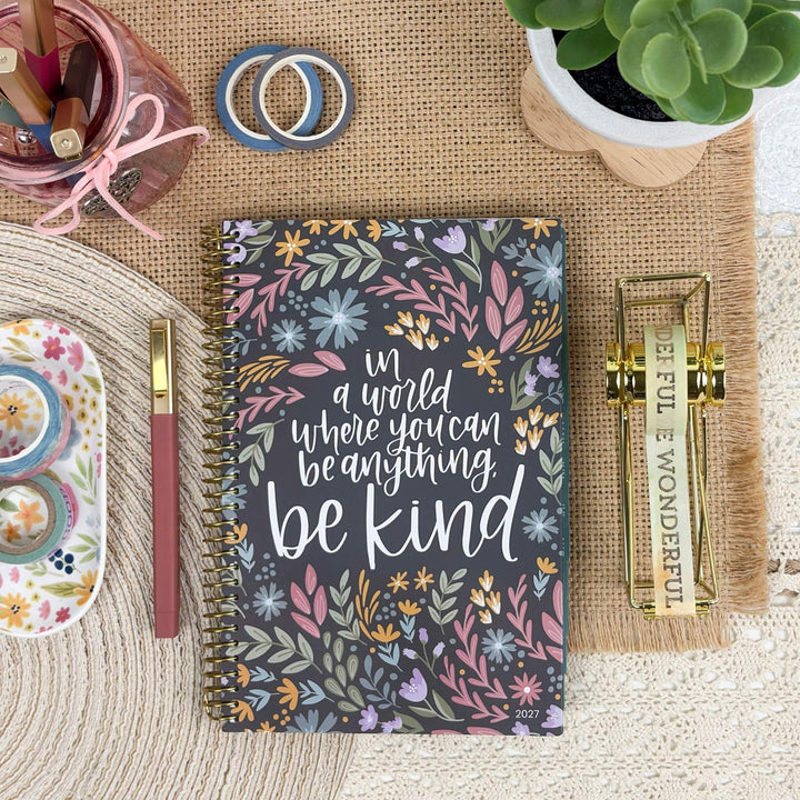 A floral spiral-bound planner or notebook with the phrase 'in a world where you can be anything be kind' in white script, styled in a flat lay on a burlap mat with stationery props including a pen, washi tapes, a small succulent, and a decorative dish.