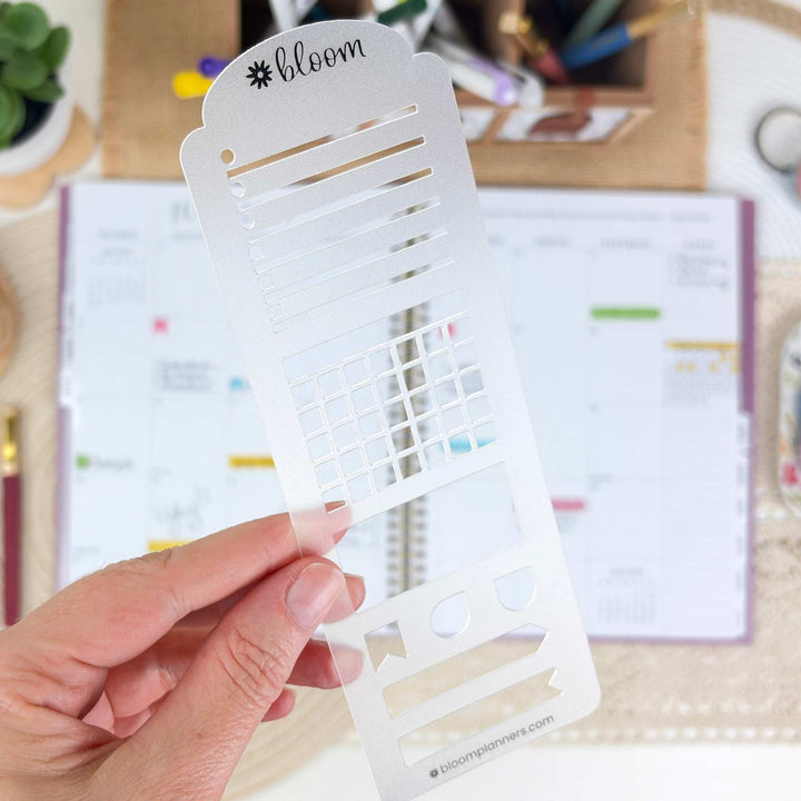 A hand holds a white plastic planner insert with multiple cutout slots and the brand name 'bloom' printed at the top, with a blurred planner in the background.