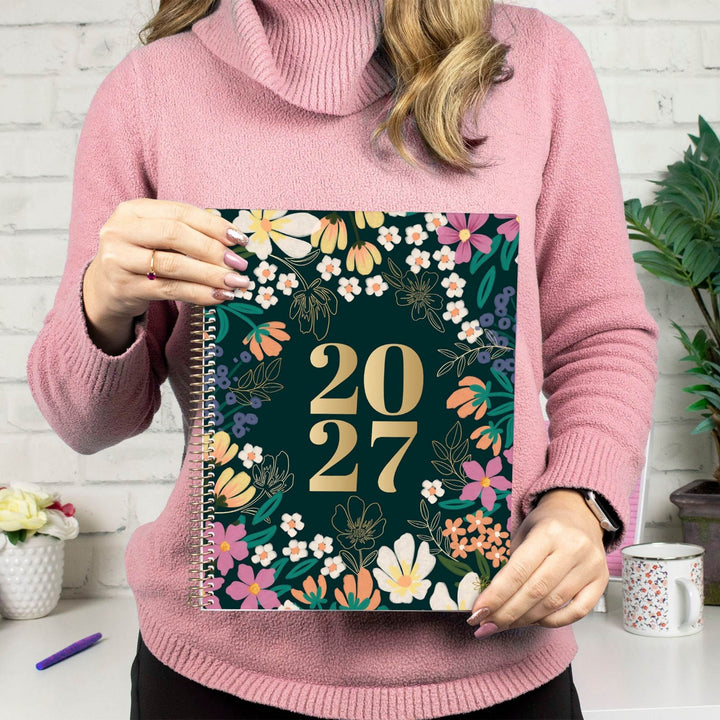 A woman in a pink turtleneck sweater holds a 2027 spiral-bound planner with a dark green floral cover in front of a white brick wall.