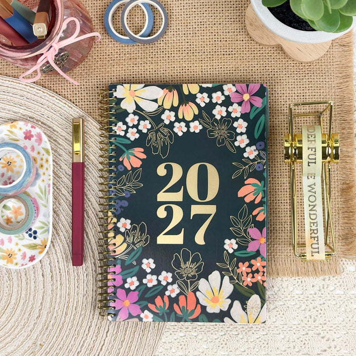 A spiral-bound 2027 planner with a dark teal floral cover is arranged on a woven mat with desk accessories around it, including a pen, washi tape, a pencil case, and a small plant.