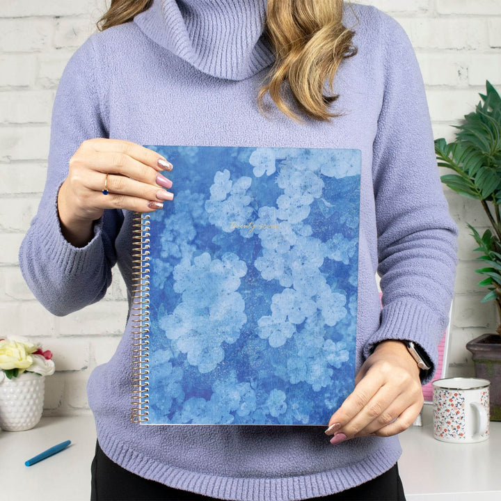 A person in a lavender turtleneck sweater holds a blue spiral notebook with a floral pattern in front of a white brick wall background.
