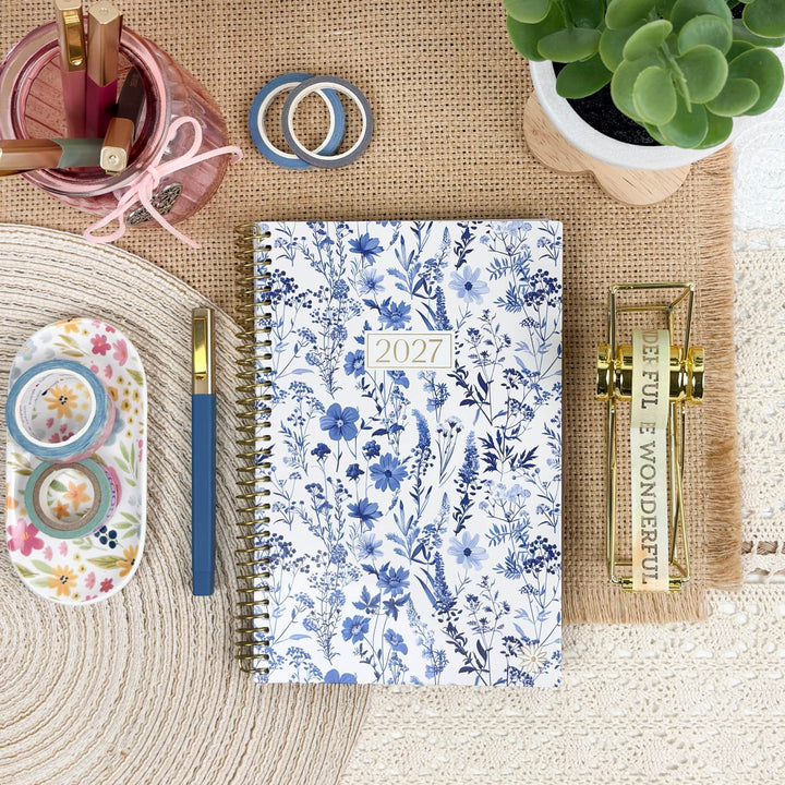 A 2027 spiral-bound planner with a blue floral pattern on a white background, styled in a flat-lay arrangement with stationery, a succulent, and decorative props on a burlap mat.